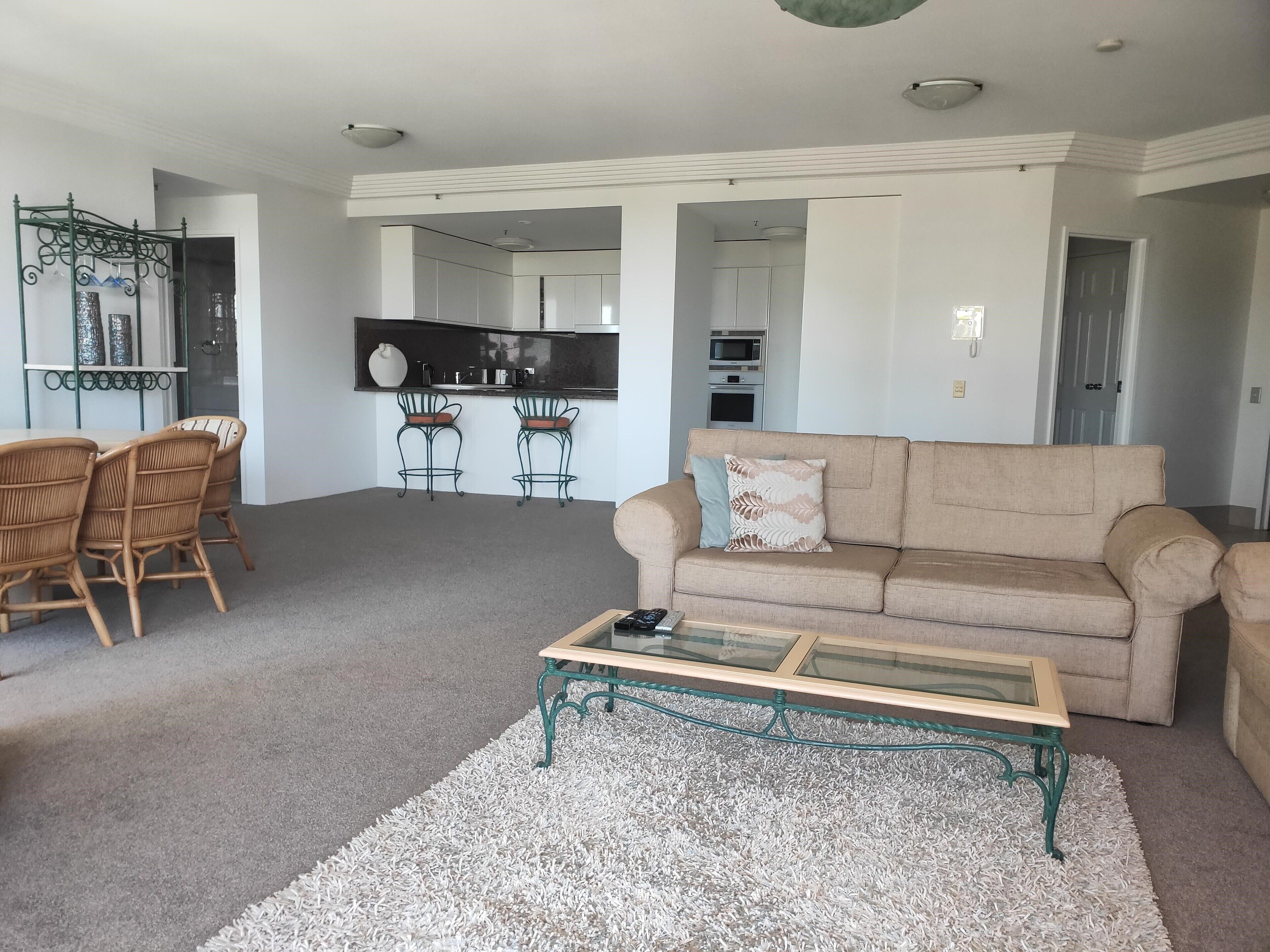 Una sala de estar con un sofá color beige, dos sillones de mimbre, una mesa de centro de vidrio y una cocina al fondo.
