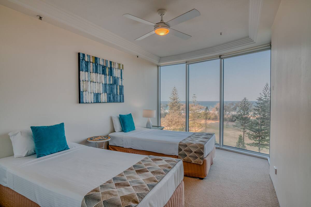 A bedroom with two beds, a ceiling fan, a large window with a view of trees, and a painting on the wall.