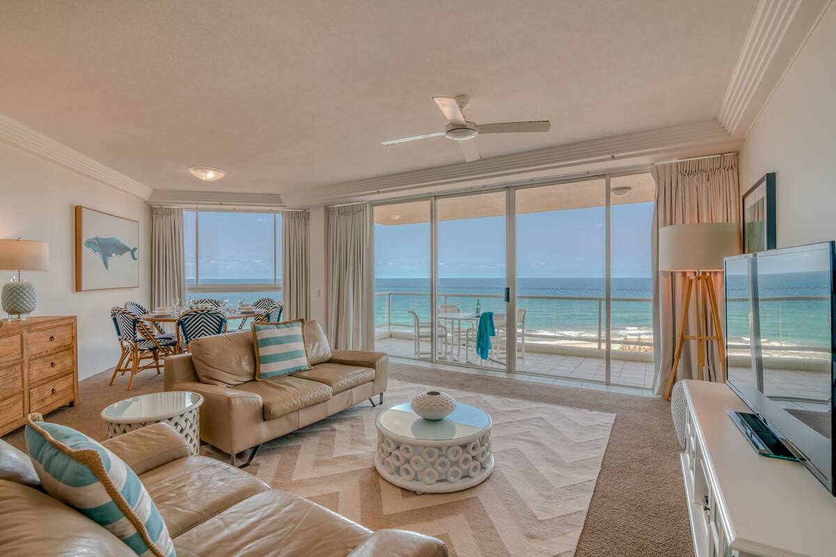 A spacious living room with a large window overlooking the beach, a ceiling fan, and a flat-screen TV.
