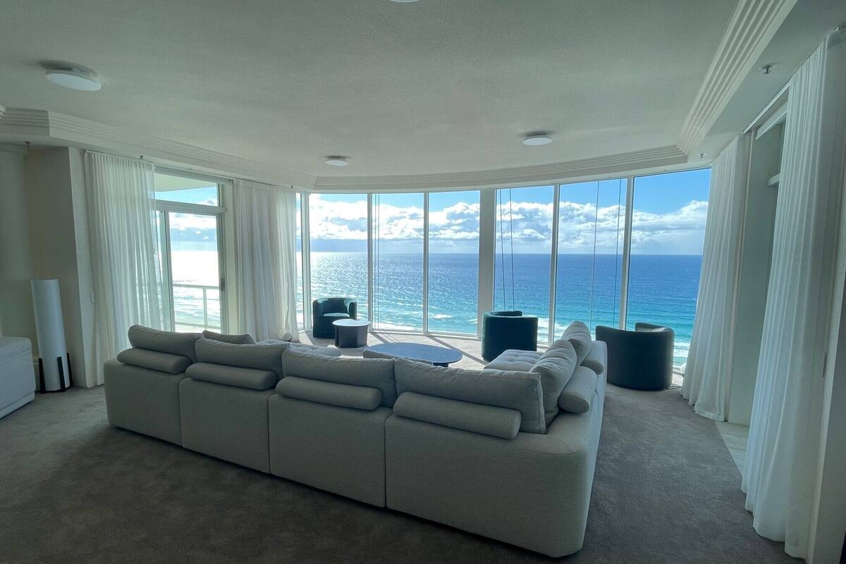 A spacious living room with a large grey sectional sofa, a small round coffee table, and a view of the ocean through large windows.