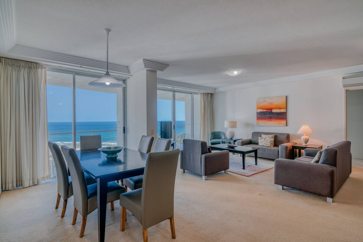 A modern hotel room with a dining area, a sofa set, and a large window with a view of the ocean.