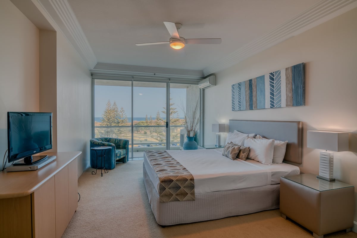 A modern bedroom with a large bed, a desk with a computer, a chair, and a view of the outdoors.