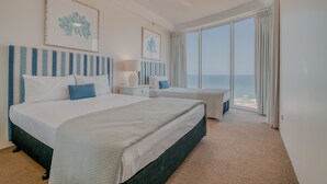Habitación de hotel con una cama grande, una cama más pequeña, una mesita de noche, una lámpara y vistas al océano.