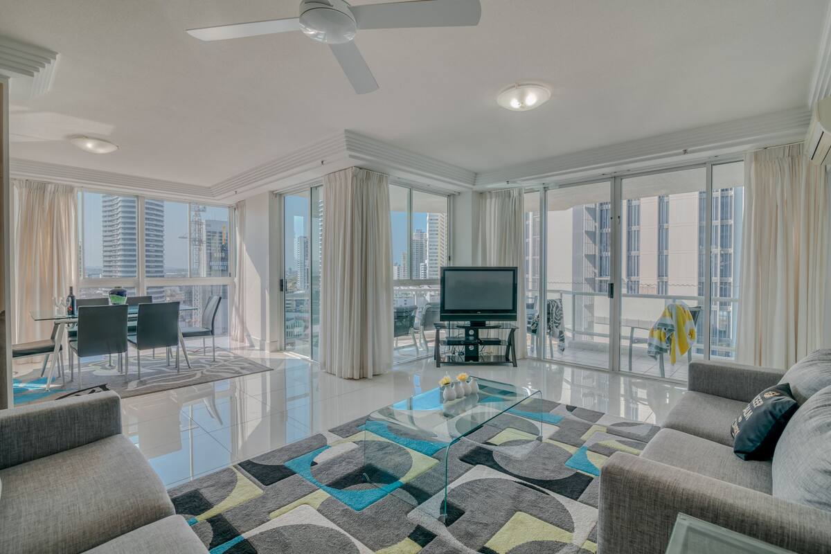 A spacious living room with a glass coffee table, a flat-screen TV, and a balcony with a view of the city.