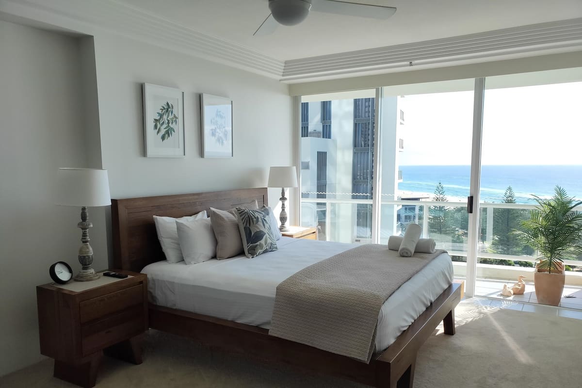 A bedroom with a large bed, a nightstand, a lamp, and a view of the ocean.