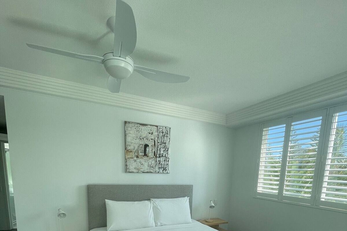 A modern hotel room with a large bed, a ceiling fan, a wall-mounted artwork, and a window with blinds.