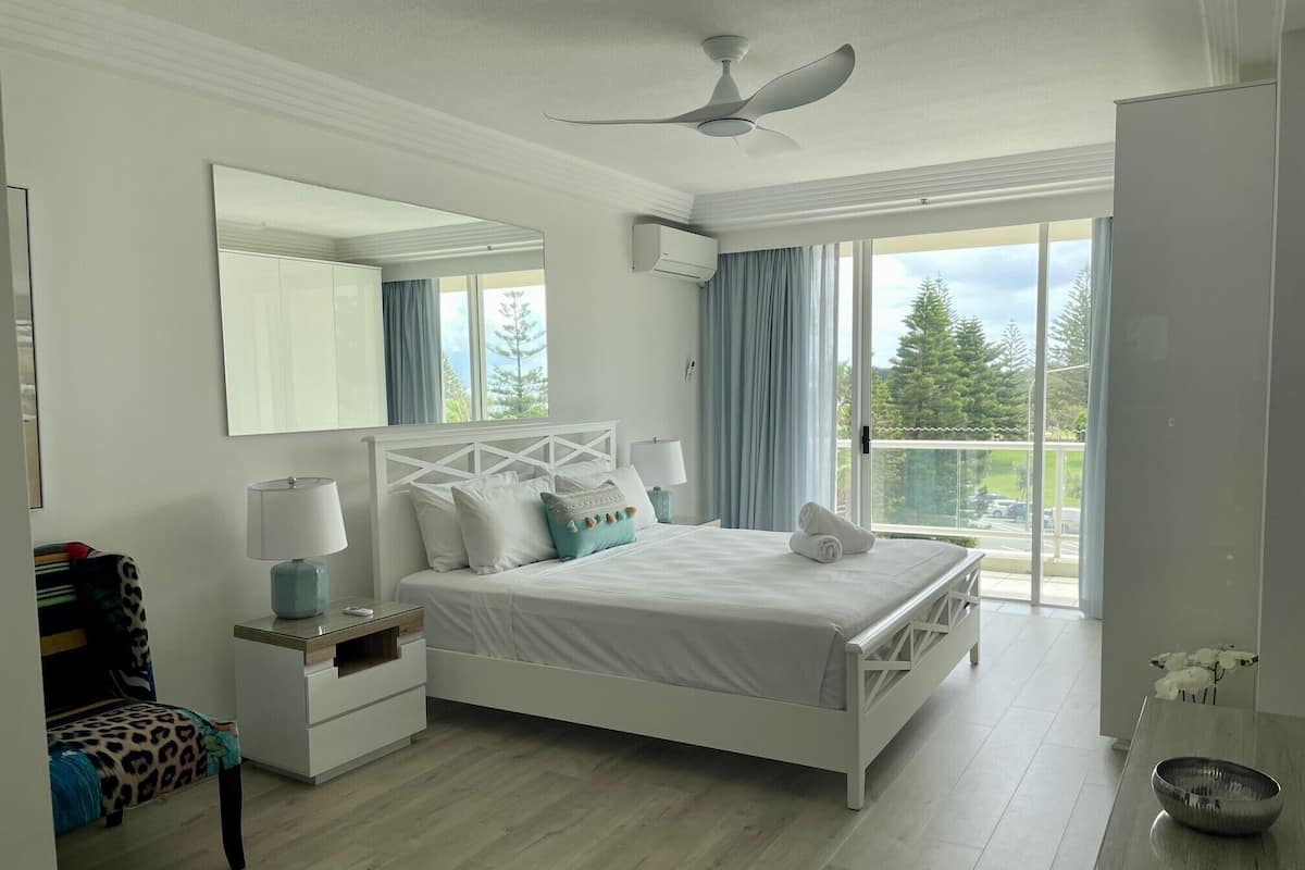 A bedroom with a bed, a nightstand, a chair, a mirror, and a view of the outdoors.