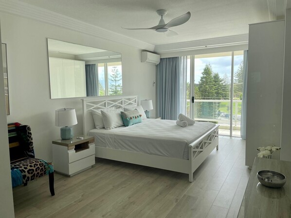 Un dormitorio con cama, mesita de noche, silla, espejo y vistas al exterior.