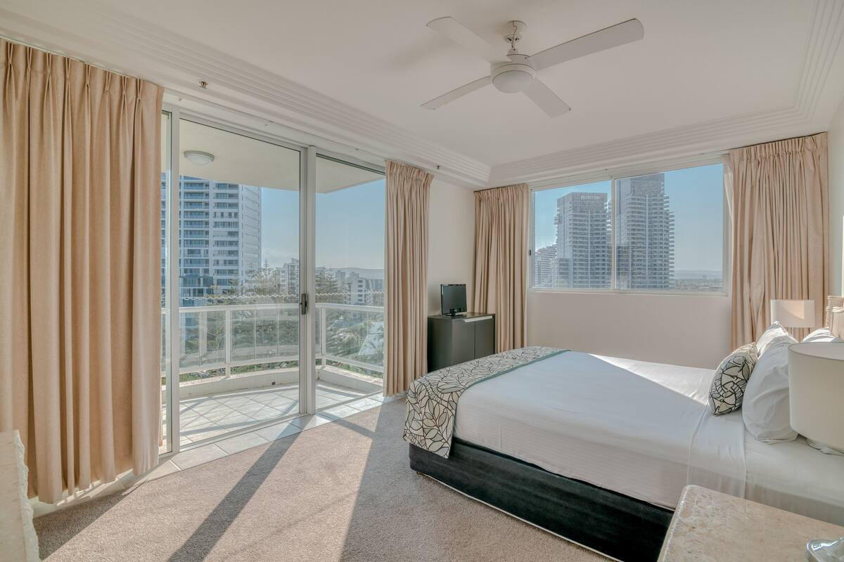 A hotel room with a large bed, a flat-screen TV, a balcony with a city view, and a ceiling fan.