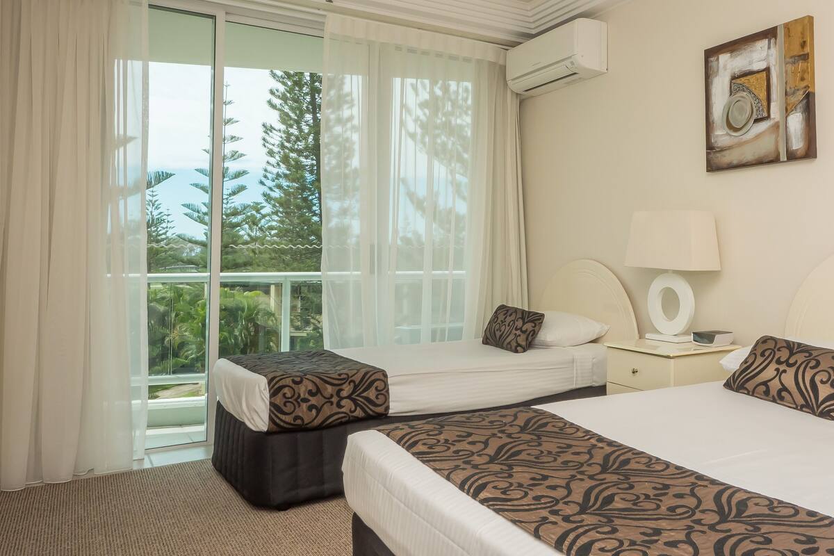 A hotel room with two beds, a window with curtains, a bedside table with a lamp, and a wall-mounted artwork.