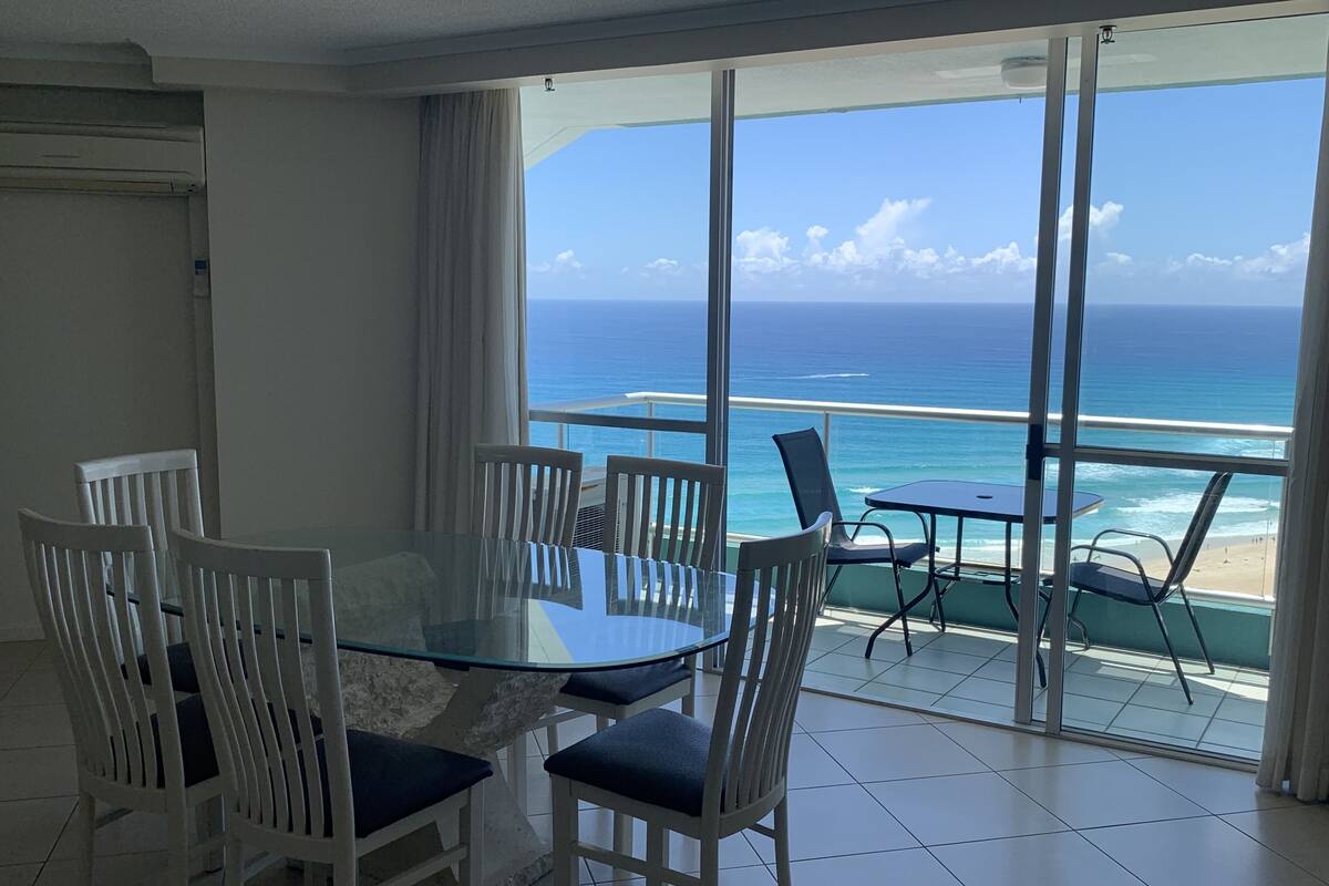 A dining area with a glass table and chairs, overlooking a beach and ocean.