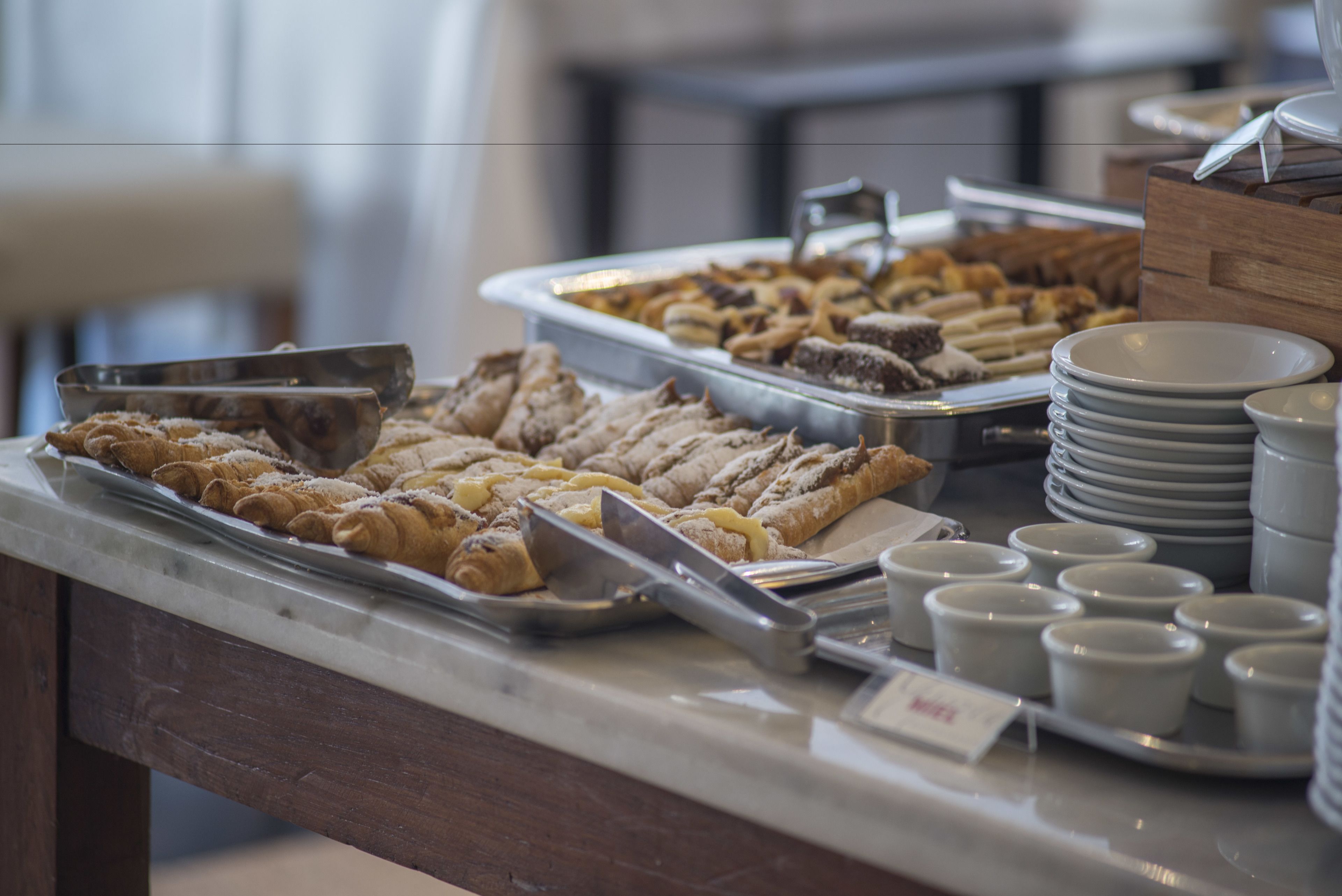 Buffet com diversos pães e sobremesas, incluindo croissants e bolos, disposto sobre um balcão de mármore com utensílios e pratos.