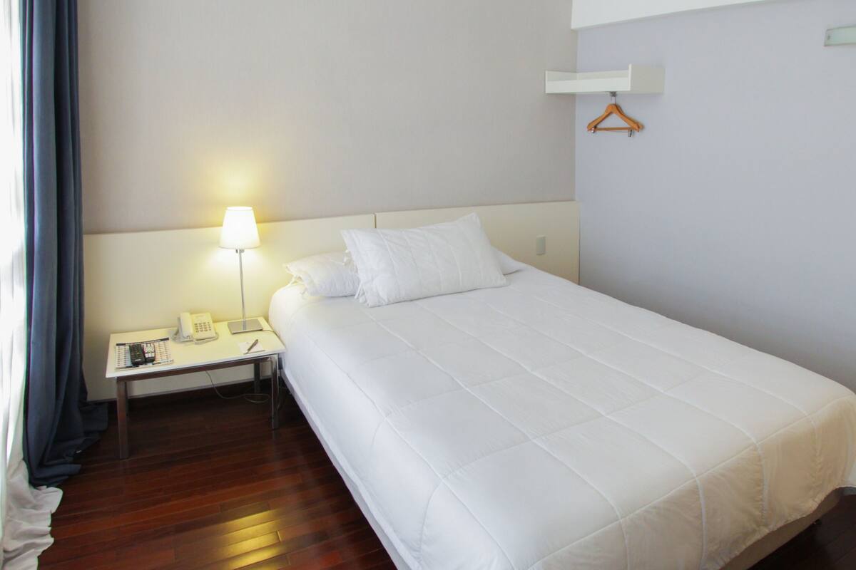 Habitación de hotel con una cama individual, una mesita de noche con lámpara, teléfono y una pequeña mesa con bolígrafo y papeles.