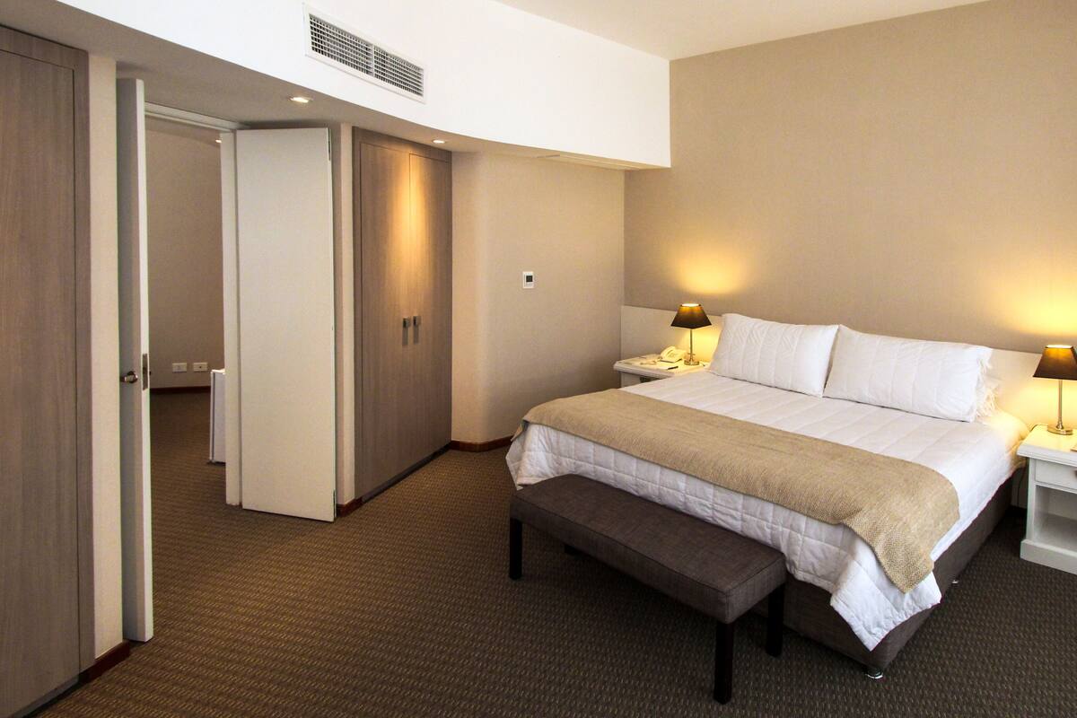 Una habitación de hotel con cama, mesitas de noche, un banco y una puerta que da a otra habitación.