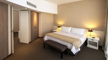 Una habitación de hotel con cama, mesitas de noche, un banco y una puerta que da a otra habitación.