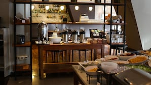 Montaje de bufé con diversos alimentos y máquinas de café, una zona de bar con botellas y una escalera al fondo.