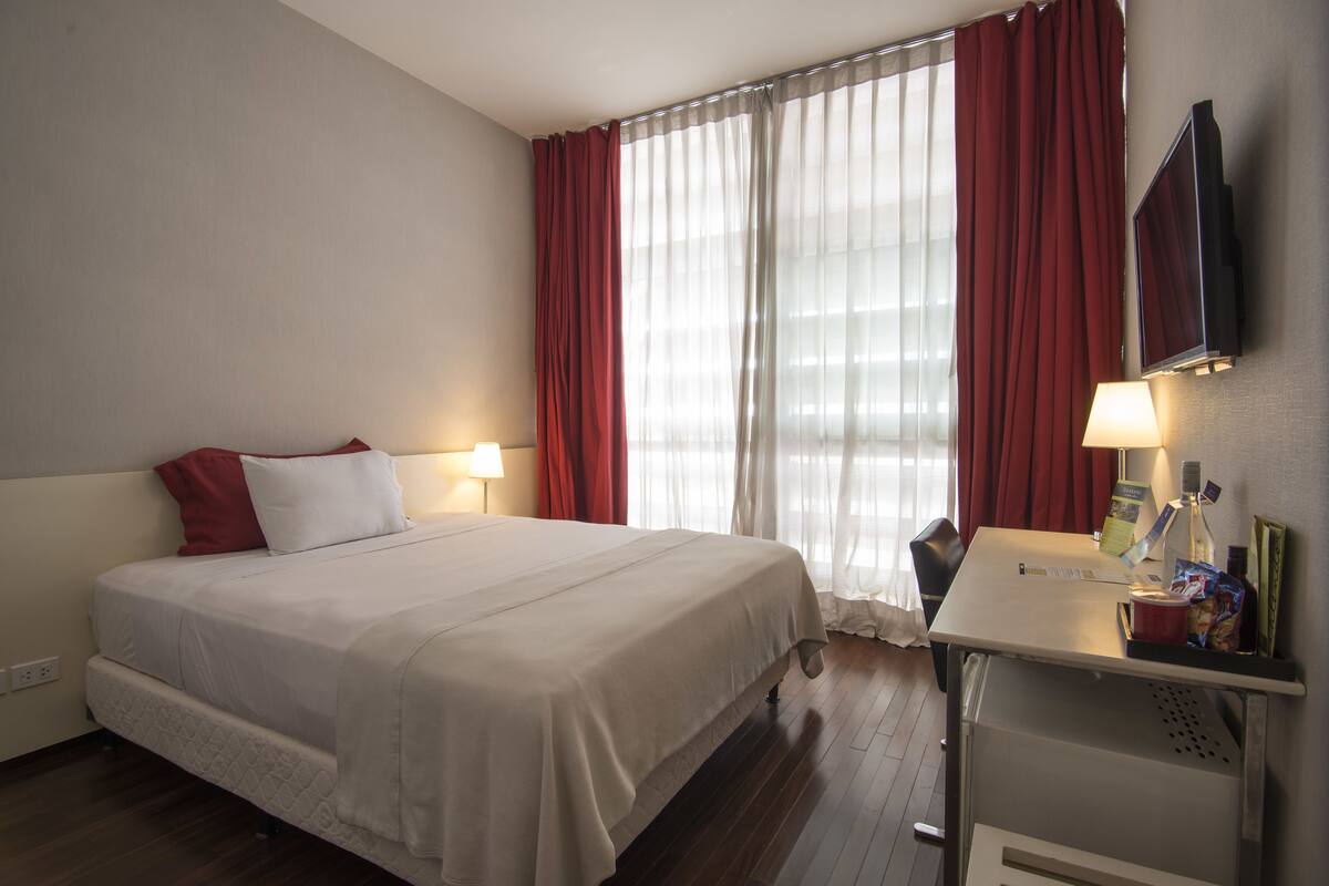 Una habitación de hotel con cama, escritorio con lámpara, televisión y ventana con cortinas.