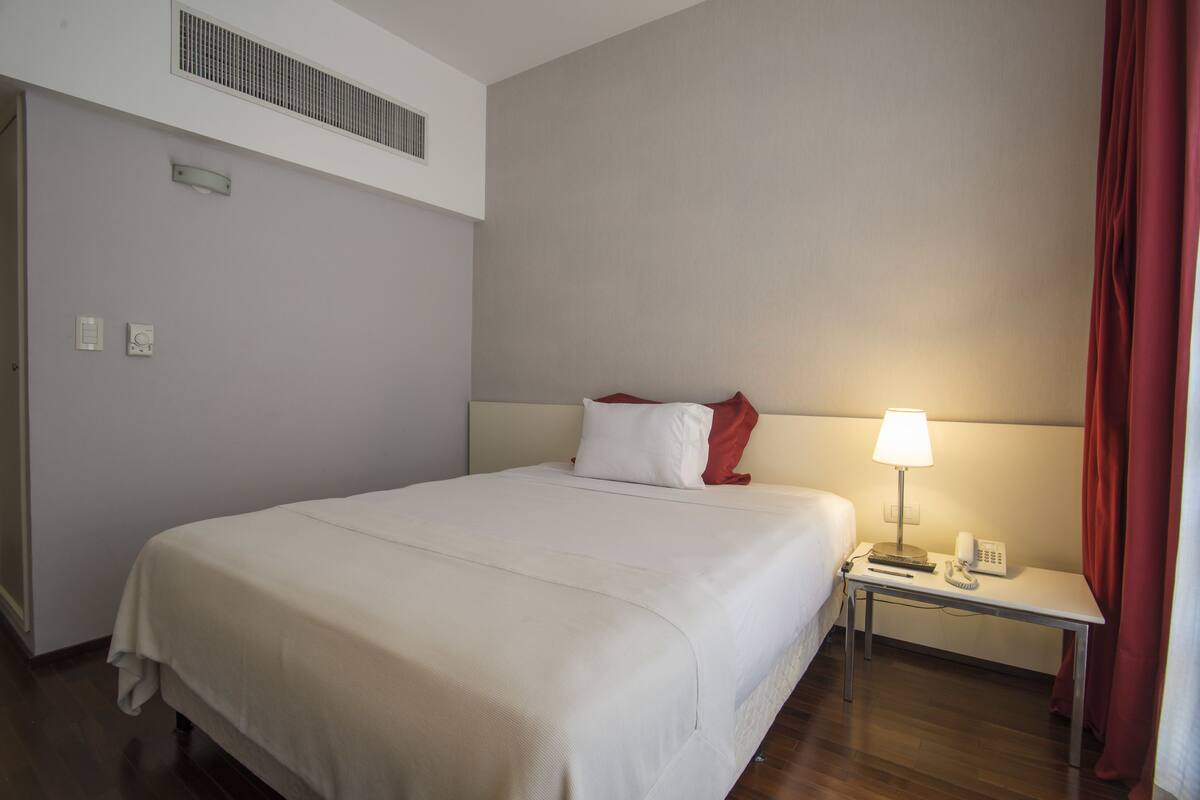Una habitación de hotel con una cama grande, una mesita de noche con lámpara, un teléfono y una pequeña mesa.