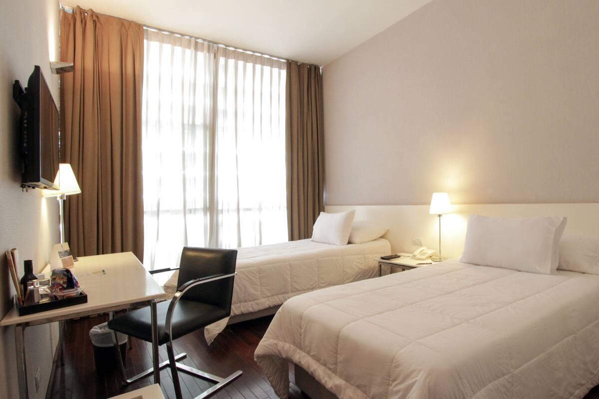 Una habitación de hotel con dos camas, un escritorio con silla, un televisor y una ventana con cortinas.