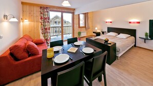 Ein modernes Hotelzimmer mit Holzboden, einem Bett mit weißer Bettwäsche, einem roten Sofa, einem Esstisch mit Tellern und Besteck sowie einem Balkon mit Holzbalkonen.