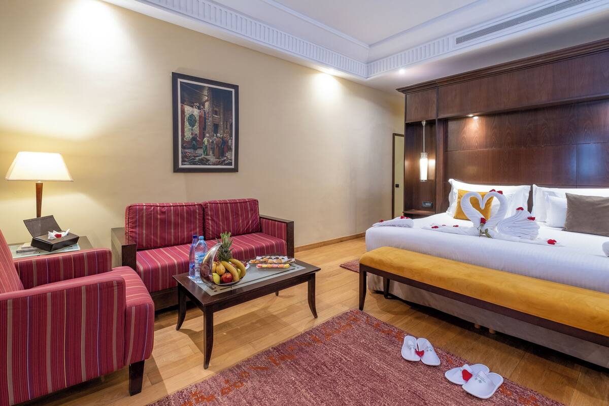 Habitación de hotel con una cama, un sofá, una mesa de centro con fruta, una lámpara y un cuadro en la pared.