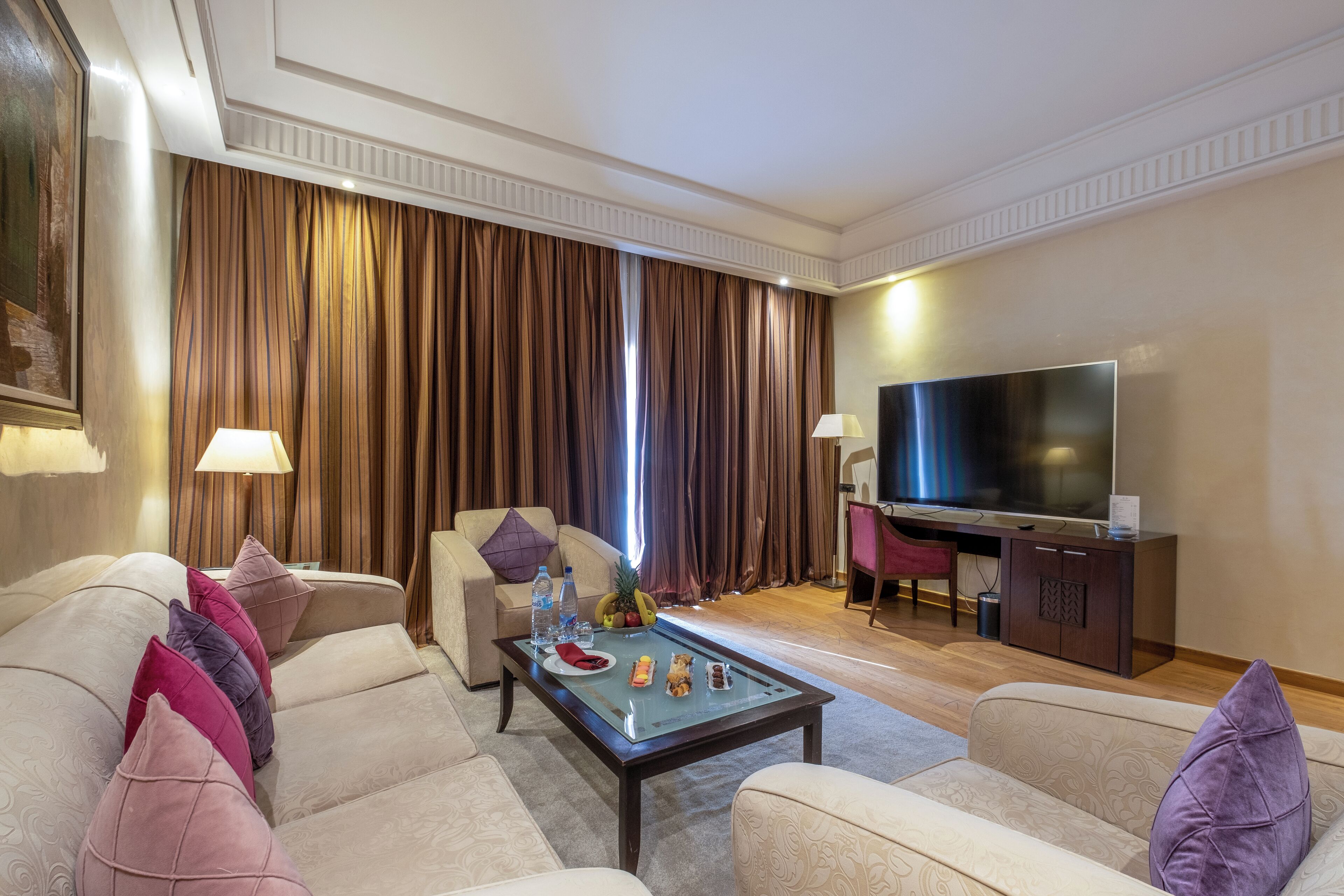 Salón de hotel con un conjunto de sofá, una mesita de centro con aperitivos, un televisor de pantalla plana y un amplio ventanal con cortinas.