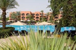 Un espace piscine d’hôtel avec des chaises longues et des parasols, entouré de palmiers et d’un bâtiment doté de balcons.