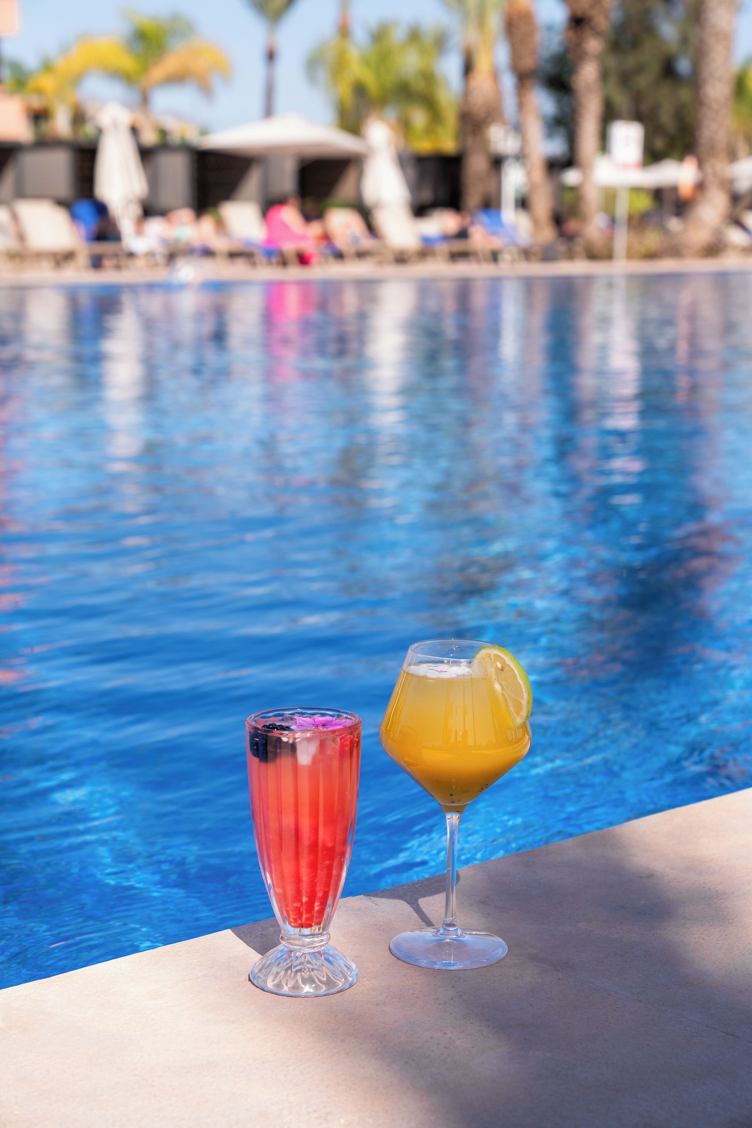 A poolside with two drinks, one red and one yellow, and a clear view of the pool.