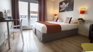 A hotel room with a bed, a desk with a chair, a lamp, and a view of the outside.