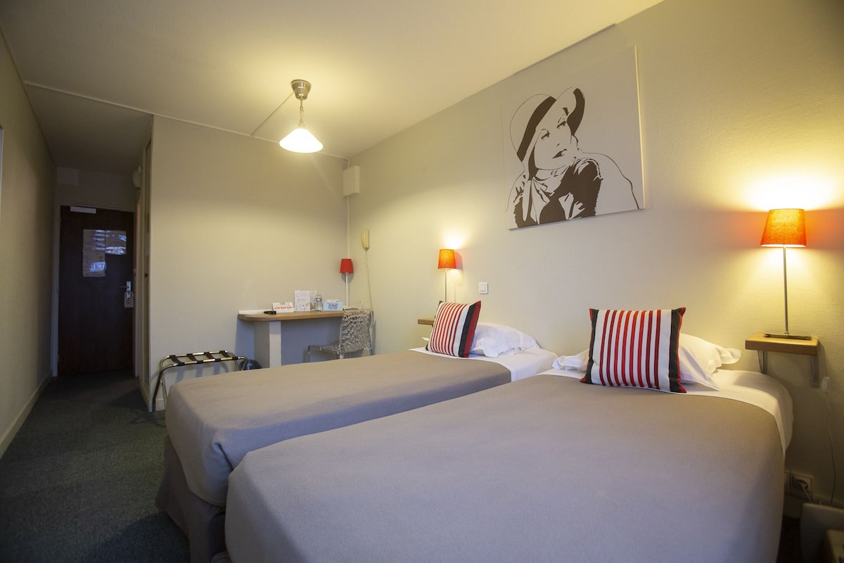 Habitación de hotel con dos camas, un escritorio y una silla. Hay lámparas a ambos lados de la cama y un cuadro en la pared.