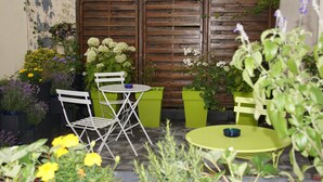 A patio with a wooden fence, two white chairs, a green table, and potted plants.