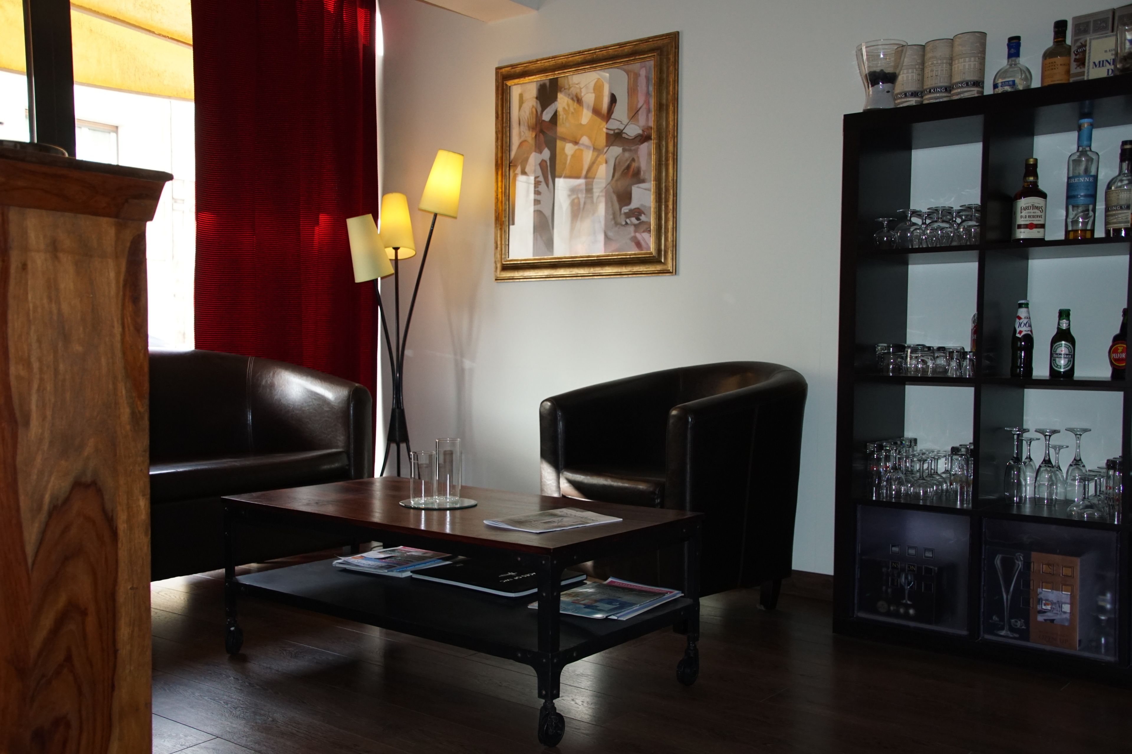 A living room with a wooden coffee table, two leather armchairs, a bookshelf with liquor bottles, and a framed abstract painting on the wall.