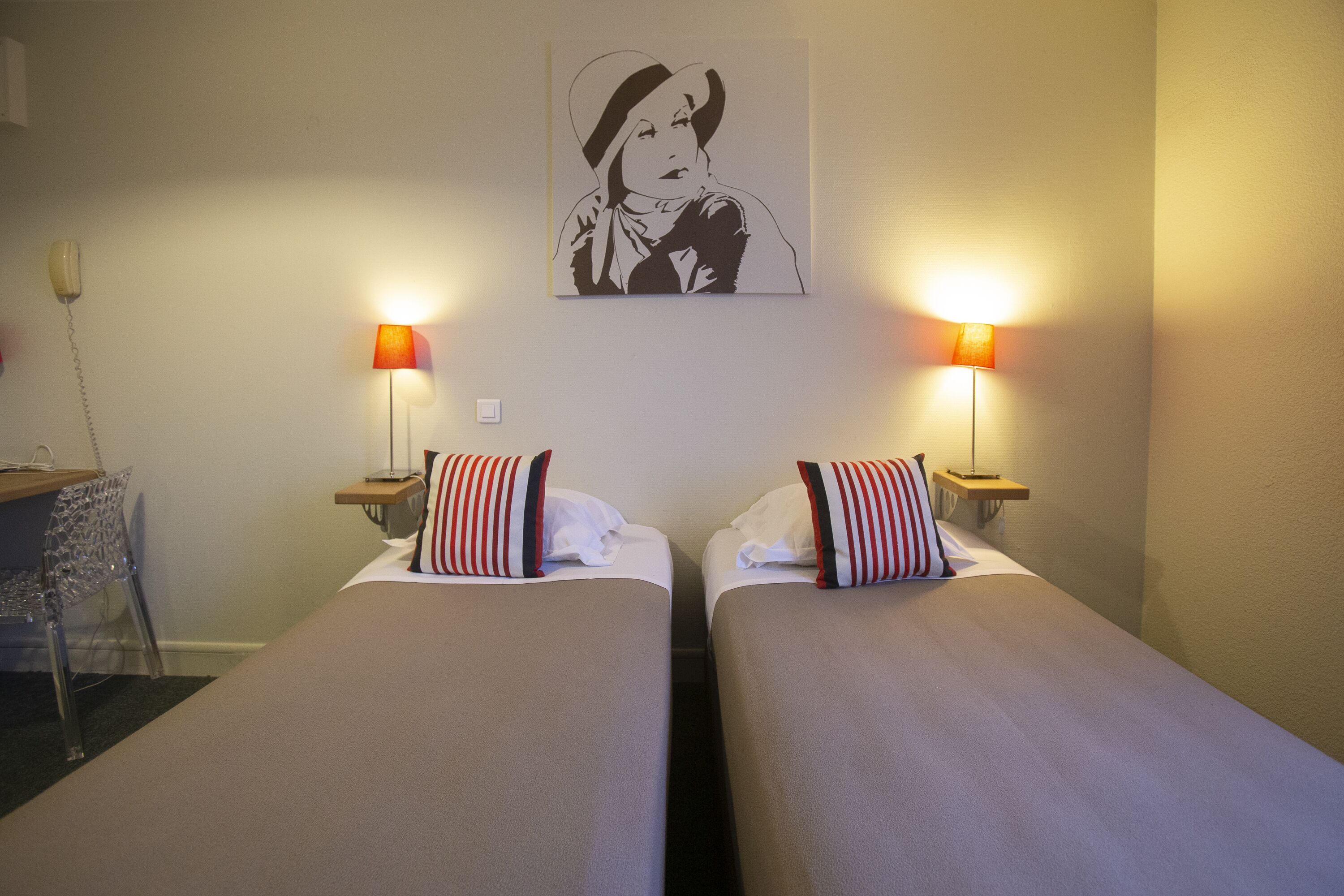 A hotel room with two single beds, each with striped pillows and a white comforter. There are two bedside lamps with orange shades, and a wall-mounted telephone. A framed artwork hangs above the beds.