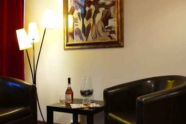 A room with a table set with a bottle of liquor, a glass, and a decorative bowl. There are two chairs and a lamp with three light bulbs. A painting hangs on the wall.