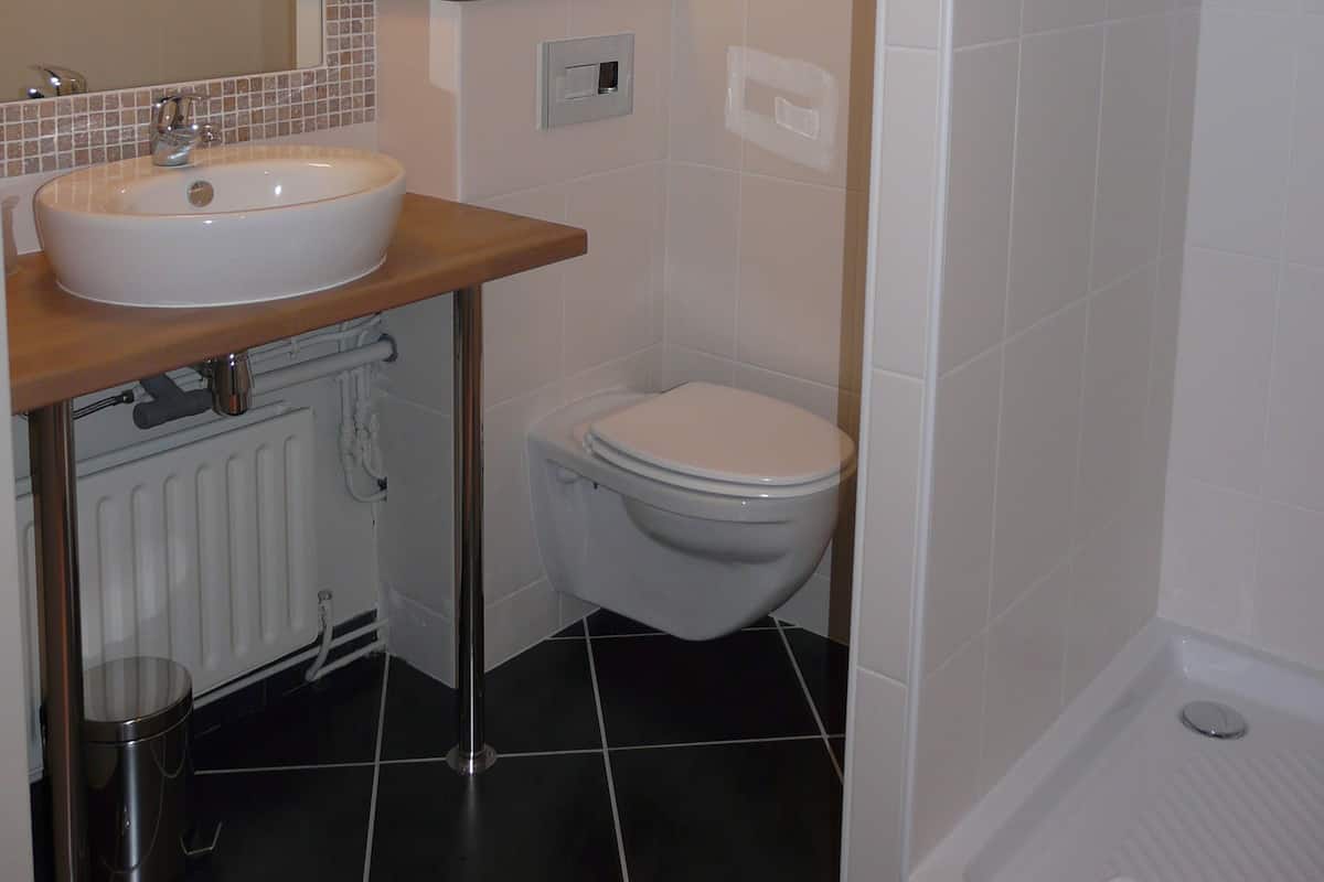 Un baño con lavamanos, inodoro y ducha blancos. Se observa un perchero con toallas y un pequeño estante sobre el inodoro.