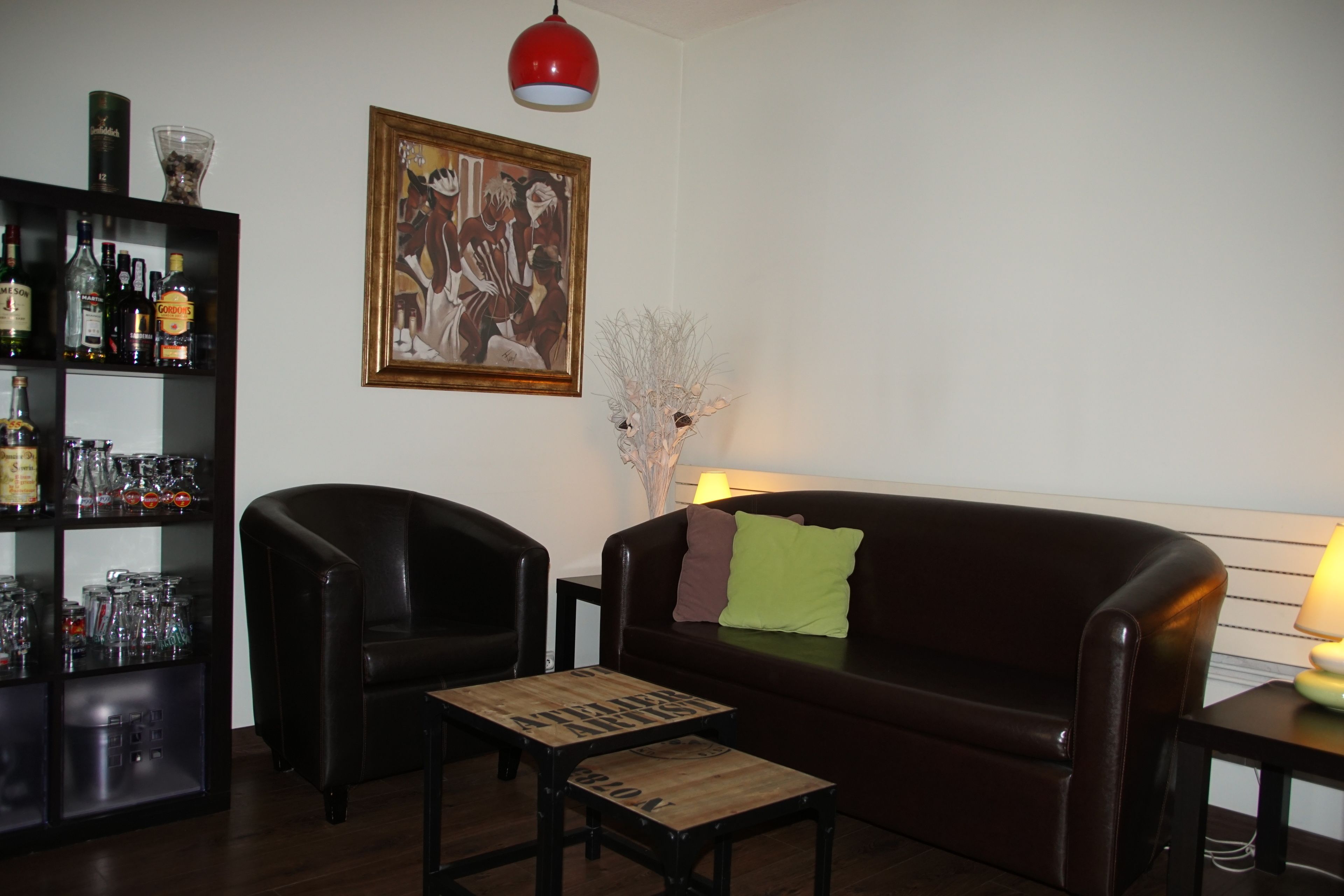 Un salon avec un canapé en cuir marron foncé, une table basse en bois, un fauteuil noir, une bibliothèque contenant diverses bouteilles et objets en verre, et un tableau encadré accroché au mur.
