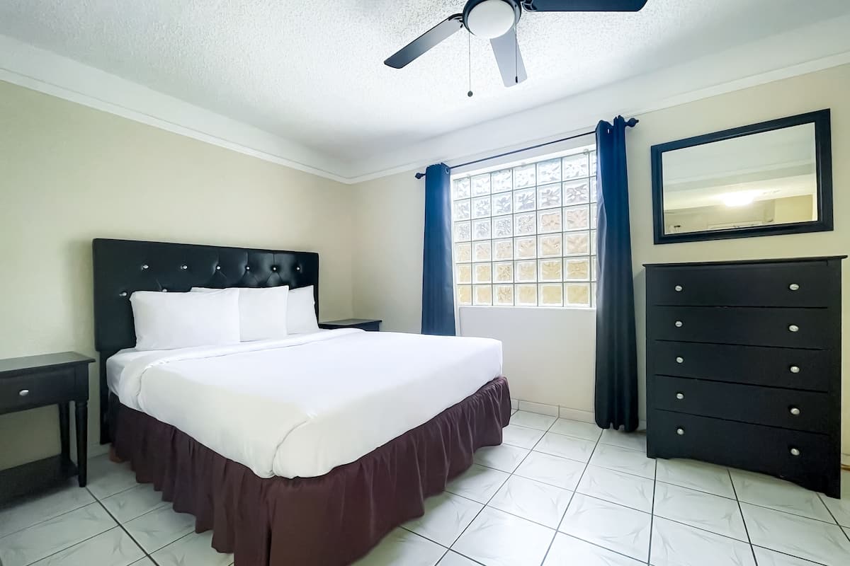 Een keurig opgemaakt slaapkamer met een bed, een dressoir, een nachtkastje, een plafondventilator en een raam met gordijnen.