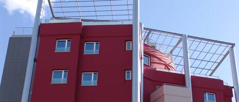 Un edificio de varios pisos con fachada roja, ventanas amplias y un elemento estructural blanco destacable.