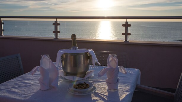 Une table dressée avec une bouteille, un bol de noix et des serviettes pliées, sur un balcon donnant sur la mer.