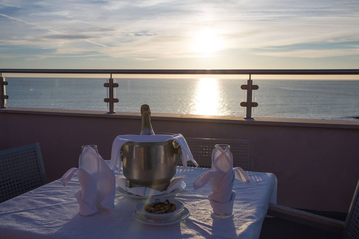 Une table dressée avec une bouteille, un bol de noix et des serviettes pliées, sur un balcon donnant sur la mer.