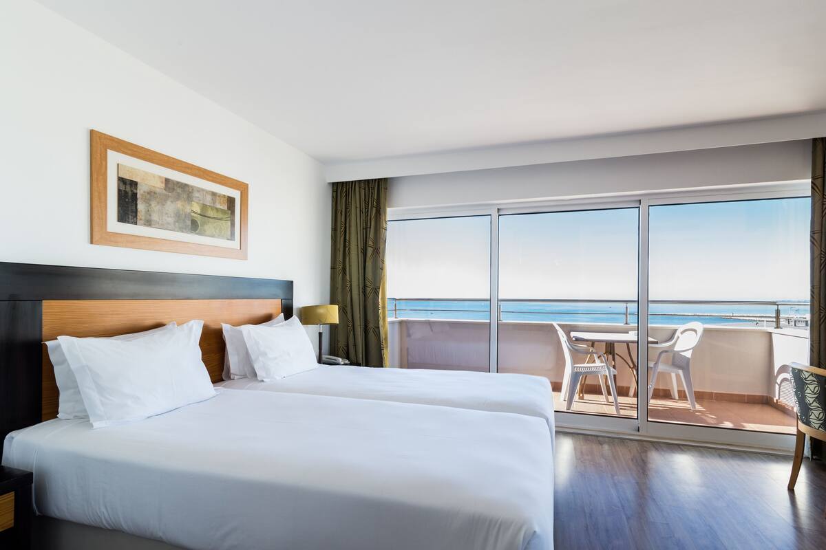 Une chambre d’hôtel avec deux lits, une grande fenêtre, un balcon avec une table et des chaises, et un tableau au mur.