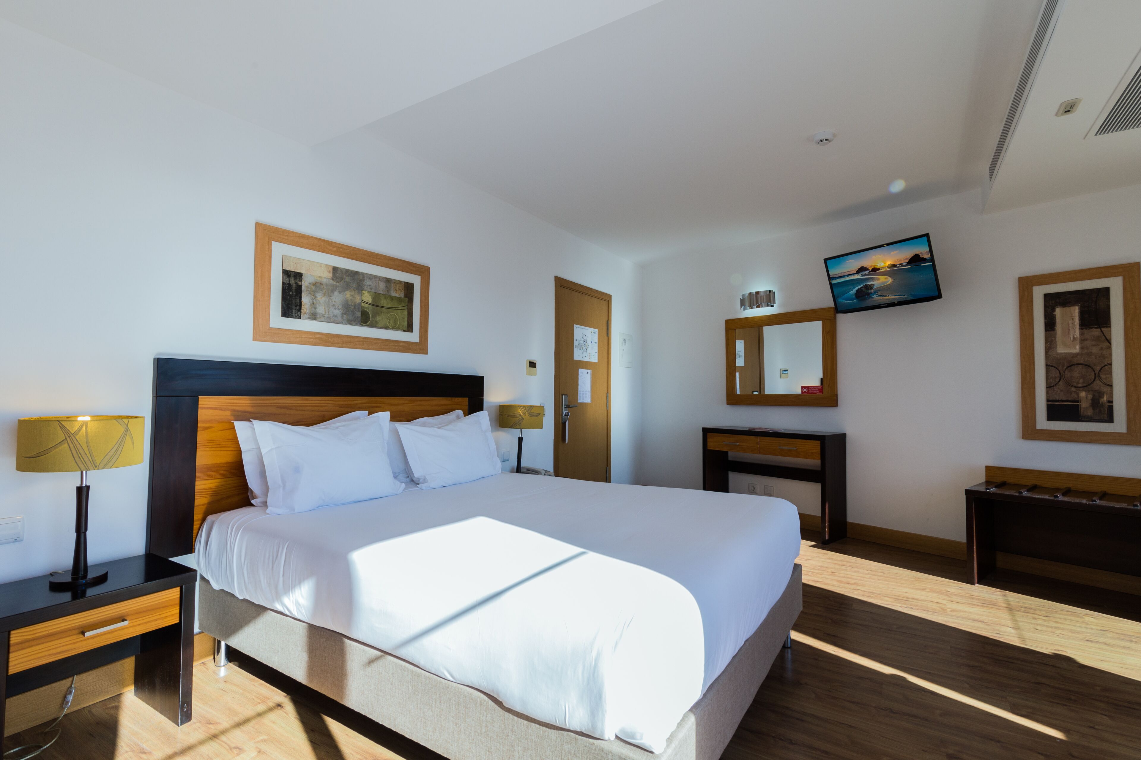 A hotel room with a large bed, bedside tables, a TV, a mirror, and artwork on the walls.