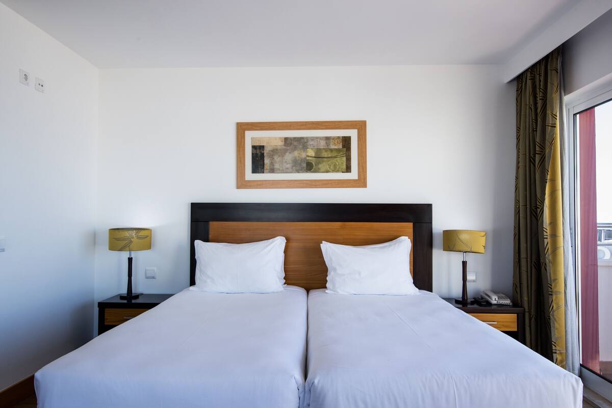 Une chambre d’hôtel avec un lit double, deux tables de chevet avec des lampes, une œuvre d’art encadrée au-dessus du lit et une fenêtre avec des rideaux.