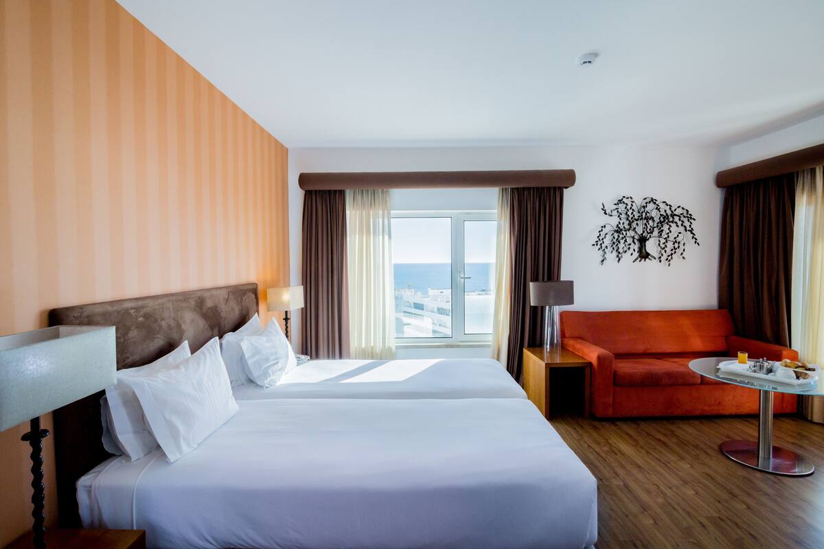 Une chambre d'hôtel avec deux lits, un canapé rouge, une petite table avec des articles pour le petit-déjeuner et une vue sur la ville.