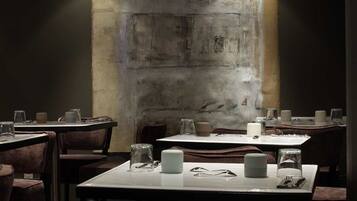 Un restaurant à la lumière tamisée, avec des tables dressées pour le dîner, caractérisé par un mur en béton rustique et des chaises en bois foncé.