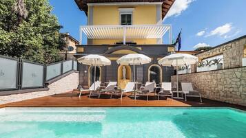 Area piscina con sedie a sdraio e ombrelloni, un muro in pietra e un edificio con un balcone.