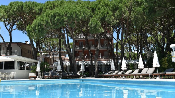 A swimming pool with lounge chairs and umbrellas, surrounded by trees and a building with balconies. - Park Hotel Maracaibo (Jesolo)