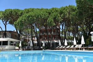 A swimming pool with lounge chairs and umbrellas, surrounded by trees and a building with balconies. - Park Hotel Maracaibo (Jesolo)