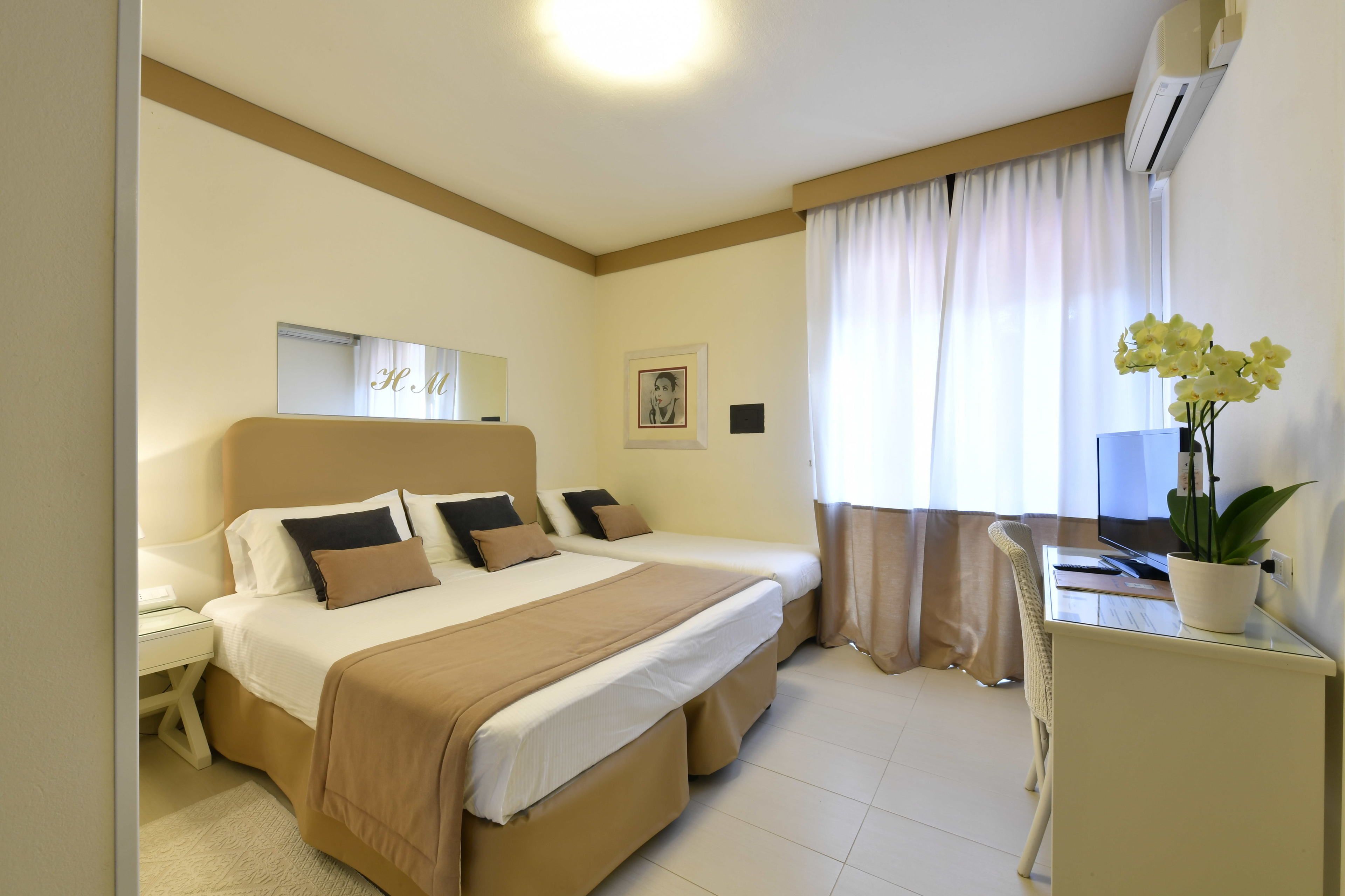 Quarto de hotel com duas camas, uma escrivaninha, uma cadeira, um vaso de plantas e uma janela com cortinas.