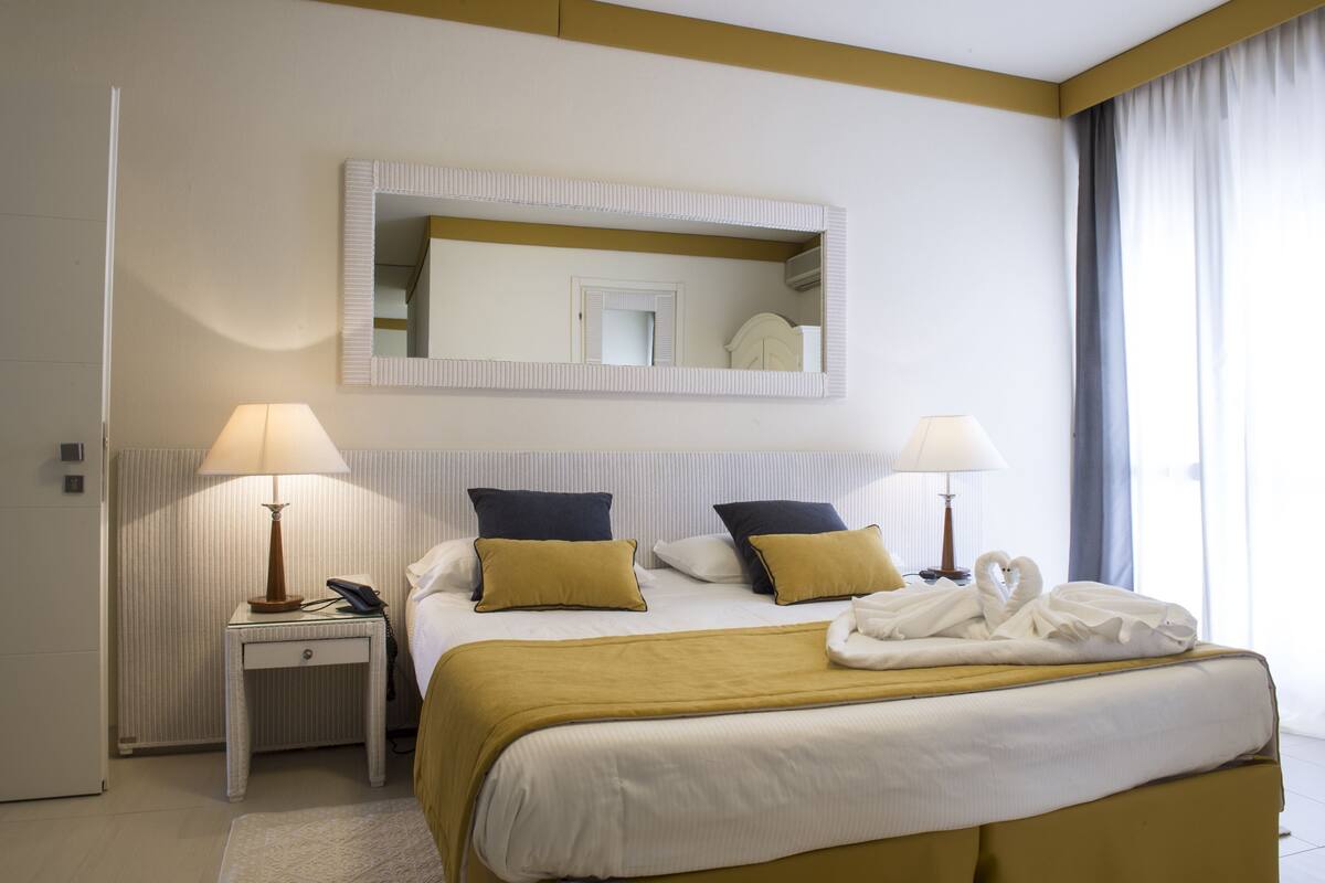 Une chambre d’hôtel avec un lit, deux lampes de chevet, une table de chevet, un miroir et une fenêtre avec des rideaux.